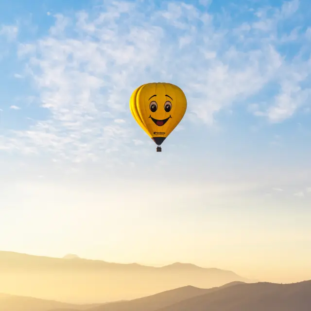 Une montgolfière jaune avec un visage de smiley volant au-dessus d'un paysage montagneux.