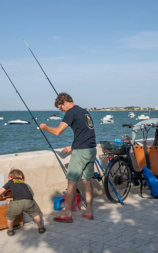Deux personnes pêchant à la ligne sur le port, entourées de bateaux amarrés.