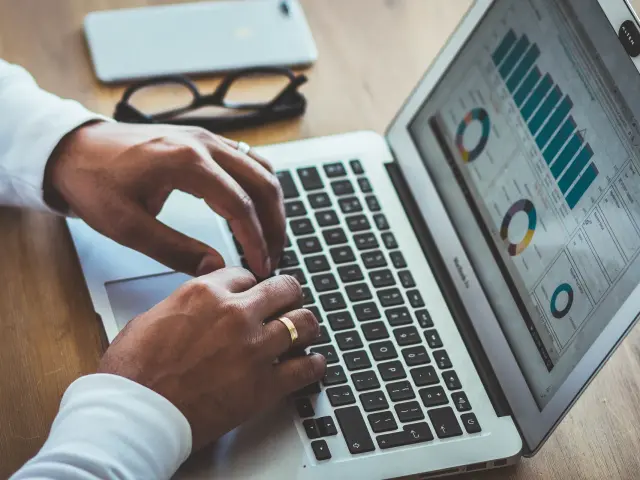 Gros plan de mains d'un homme tapant sur le clavier d'un ordinateur portable affichant des graphiques de données colorés sur un bureau en bois.