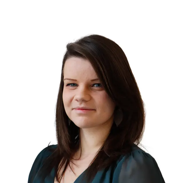 Portrait professionnel de Laura Gay, une femme souriante aux cheveux châtains longs, portant un vêtement noir, sur un fond blanc neutre.