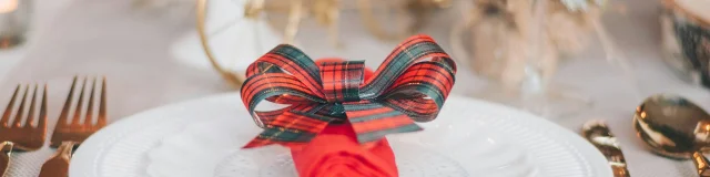 Assiette blanche ornée d’une serviette rouge roulée et nouée avec un ruban à carreaux rouges et verts.