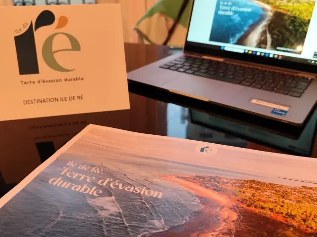Au premier plan, un dossier de presse et une carte de visite avec le logo Île de Ré, Terre d’évasion durable. En arrière-plan, un ordinateur portable ouvert sur le site web de la destination dans un décor de bureau.