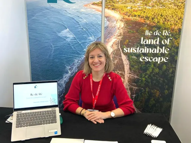 Une femme souriante en pull rouge assise derrière un stand à l'IMM de Paris. En arrière-plan, un grand panneau affiche une vue aérienne de l'Île de Ré avec le slogan land of sustainable escape. Sur la table, un ordinateur et des guides touristiques.