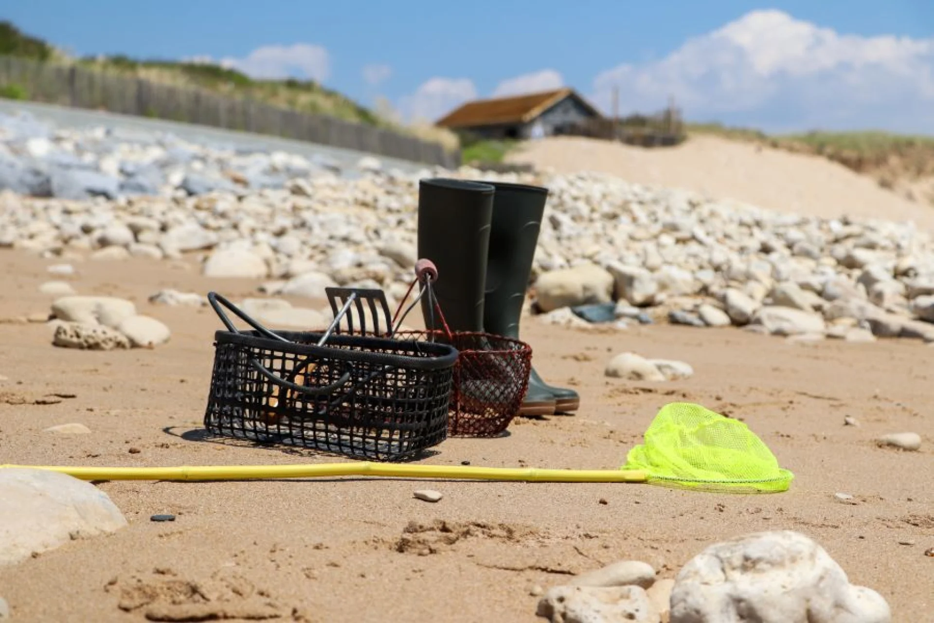 Panier, bottes et outils de pêche à pied posés sur le sable.