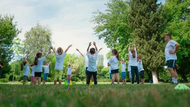 Un groupe d’enfants lève les bras pendant un exercice sportif dans un parc, encadré par un adulte.