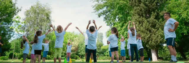 Un groupe d’enfants lève les bras pendant un exercice sportif dans un parc, encadré par un adulte.