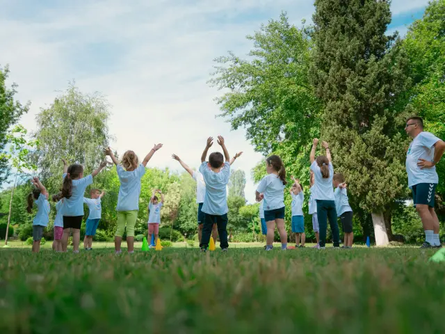 Un groupe d’enfants lève les bras pendant un exercice sportif dans un parc, encadré par un adulte.