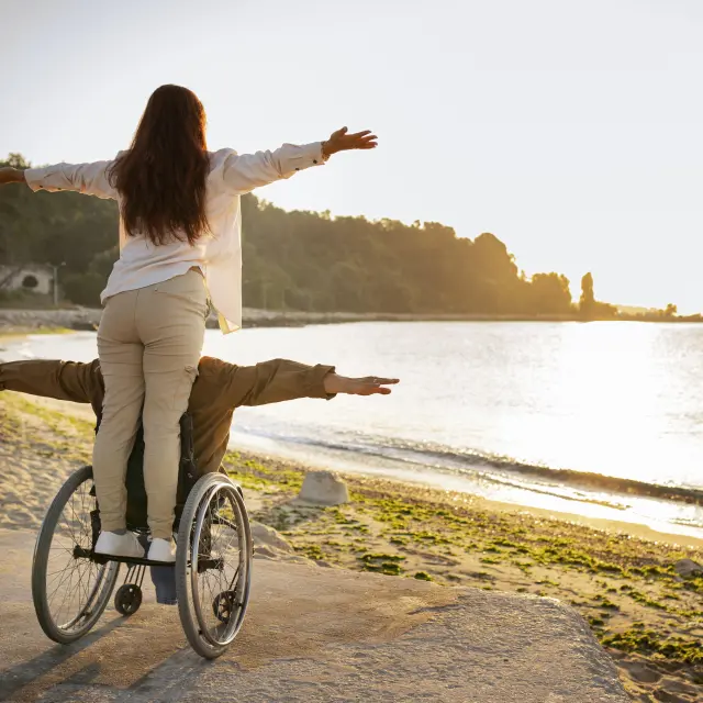 Deux personnes dont une en fauteuil roulant, bras ouverts face à la mer au coucher du soleil.